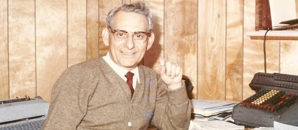 Archival photo: smiling man at a wood-paneled office desk with adding machines